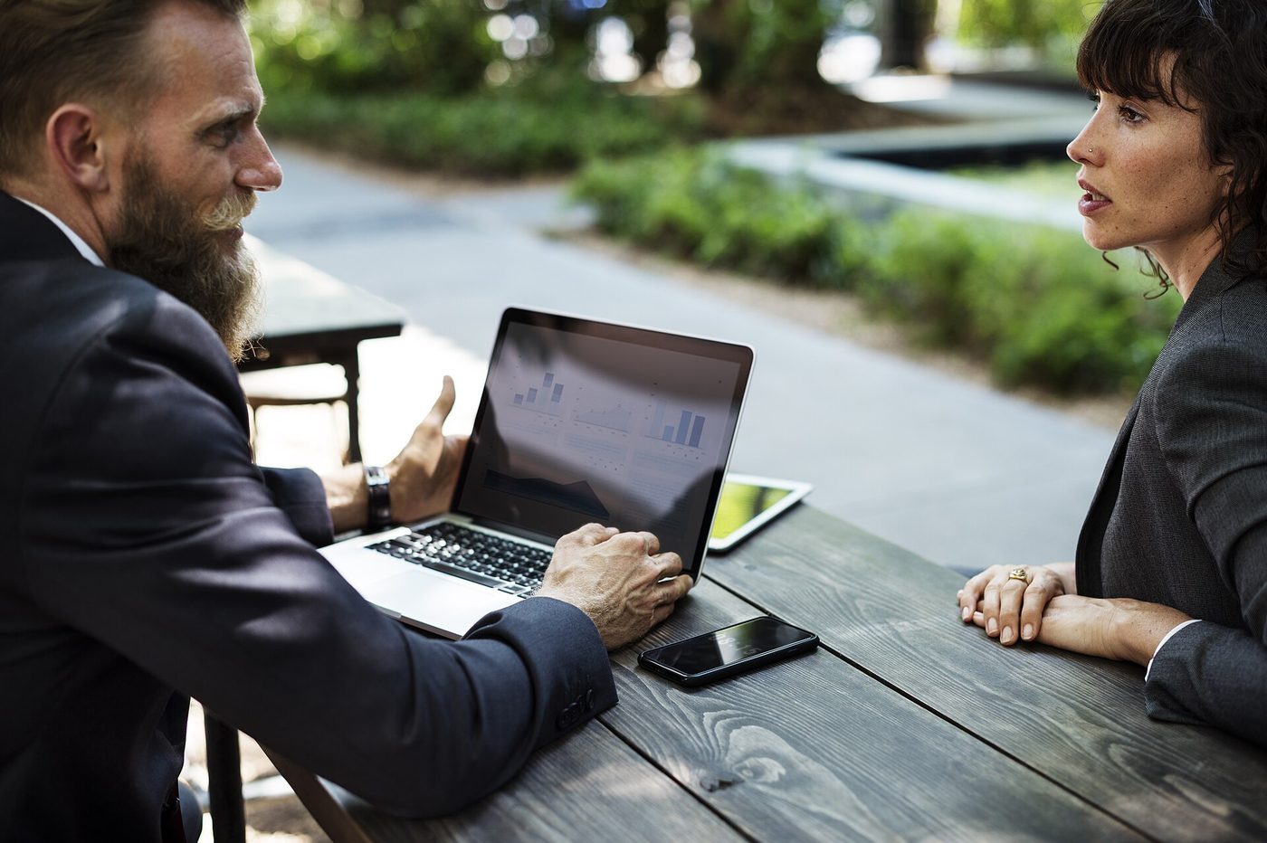 Two colleagues at an outdoor table reviewing charts on a laptop, with a smartphone and tablet beside it